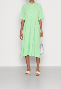 Vaaleanvihreä, lyhythihainen mekko, jossa on napitettu etuosa ja liehuva, polvipituinen hame. Yhdistetty vaaleanbeigeihin lohkokorkoisiin sandaaleihin ja laventelinväriseen laukkuun.