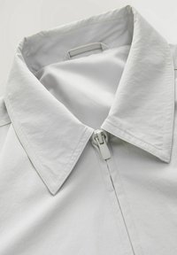 Lichtgrijze jas met een grote kraag, een gladde textuur en een ritssluiting aan de voorkant. Subtiele stikseldetails langs de randen.