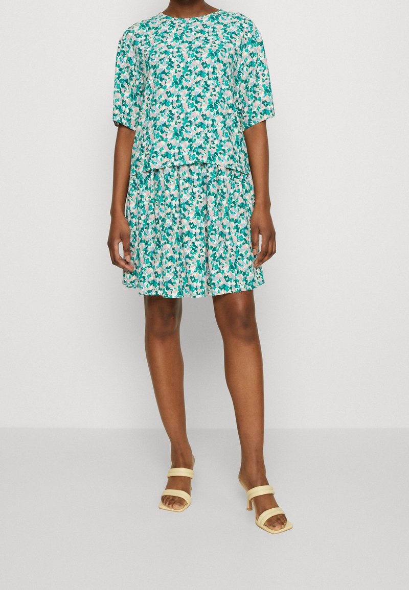 Vestido floral en verde azulado con patrones en rosa y verde. Mangas cortas, escote redondo y falda con volantes. Combinado con sandalias de tacón amarillas claras.