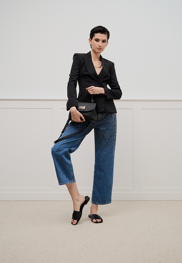 Blazer noir ajusté, pantalon en denim bleu large et sandales noires à glissière. La tenue est accompagnée d'un petit sac à main noir avec un logo métallique.
