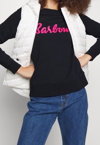 Gilet rembourré blanc sur un sweat noir avec un texte brodé rose. Associé à un jean en denim bleu, montrant une tenue décontractée.