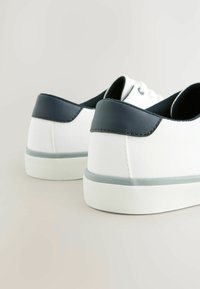 Witte canvas sneakers met een marineblauw leren hielkap, voorzien van een gestructureerde witte zool en subtiele grijze accenten langs de onderrand.