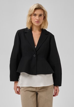 Frau mit schulterlangem blondem Haar trägt einen schwarzen Blazer über einer weißen Bluse und beigen Hosen, stehend vor einem schlichten, hellen Hintergrund.