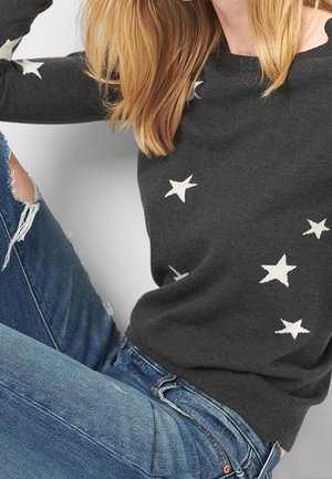 Vrouw die een zwarte trui met witte sterpatronen draagt en versleten blauwe jeans, zittend met één knie omhoog.