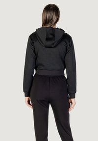 Felpa nera con un motivo testurizzato e cappuccio. Abbinata a pantaloni neri coordinati, che mostrano una vita aderente e maniche a polsino.