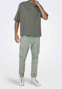 Olive green oversize t-shirt połączony z jasnoskalnym spodniami cargo z bocznymi kieszeniami i elastycznymi mankietami, noszonymi z białymi trampkami.