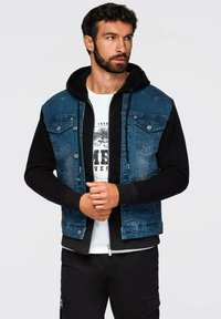 Gilet di jeans con gradiente blu scuro, bottoni a pressione e due tasche sul petto, indossato sopra un felpa nera con cappuccio e una t-shirt bianca con grafica.