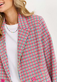 Žena oblečená v barevném blejzru s růžovými, modrými a žlutými kostkami přes bílý top, s korálkovým vícebarevným náhrdelníkem, se usmívá.