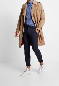 Beige trenchcoat over een blauw patroonshemden, marineblauwe slim-fit broek en witte sneakers. De jas heeft een dubbelgeknoopte stijl en zakken.