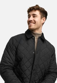 Chaqueta negra acolchada con cuello de pana, cierre a presión y superficie texturada. Llevada sobre un suéter de cuello alto beige claro.