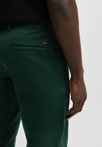 Pantaloni verde închis cu o textură netedă, având un buzunar lateral și un mic logo portocaliu "BOSS". Prezentat din lateral, cu o parte a mâinii vizibilă.