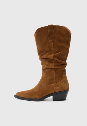 Botte de cowboy en suede marron avec un design décontracté, un bout pointu et un talon empilé. Elle présente des accents de couture et une bride de traction à l'arrière.
