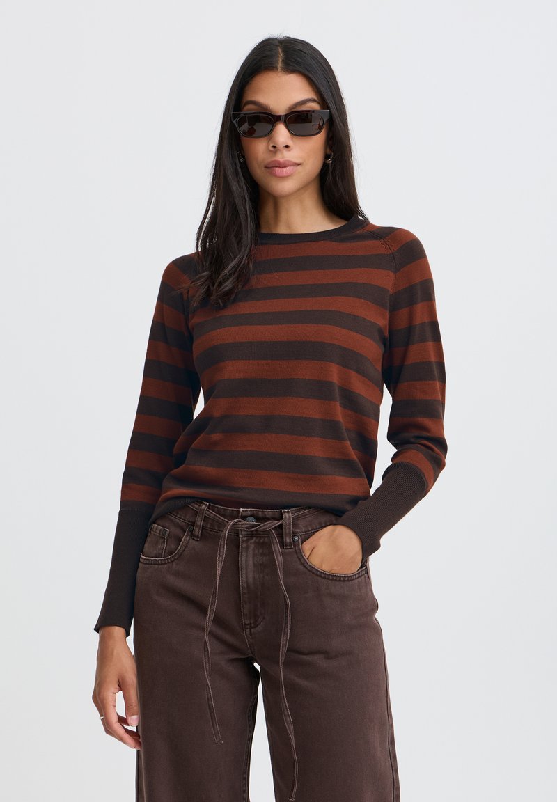 Femme portant des lunettes de soleil foncées, un haut à manches longues rayé marron et noir, et un pantalon taille haute marron foncé avec un cordon noué.