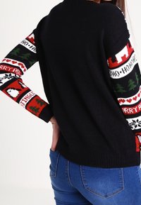 Svart stickad tröja med färgglada mönstrade ärmar som har röda, vita och gröna motiv, inklusive träd och festliga motiv.