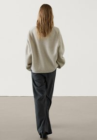 Femme aux cheveux ondulés blonds portant un pull à côtes beige ample et un pantalon large à rayures gris foncé, s'éloignant sur un sol uni.