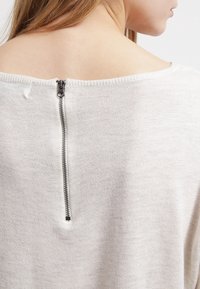 Ljusgrå tröja med bred halsringning, stickad textur och en synlig dragkedja i ryggen med metalltanddetalj.