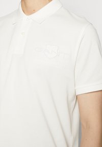 Polo shirt blanc avec une texture douce, doté d'une patte de boutonnage à trois boutons et d'un logo en relief sur la poitrine gauche. Manches courtes, coupe classique.