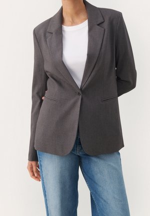 Femme portant un blazer gris foncé à un seul bouton sur une chemise blanche côtelée, associée à un jean denim bleu clair, sur un fond uni.