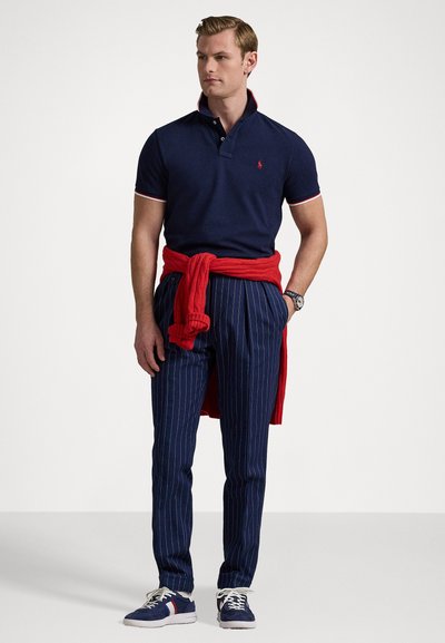 Polo navy com detalhes em vermelho e branco, combinado com calças navy risca de giz. Suéter vermelho atado à cintura, ténis azul e branco.