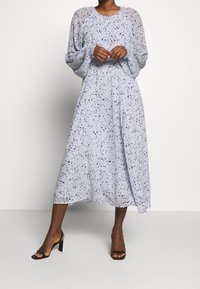 Hellblaues, langärmliges Midikleid mit lockerer Passform, das ein gepunktetes schwarzes Muster und geraffte Ärmel aufweist. Kombiniert mit schwarzen High-Heels.