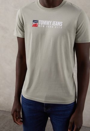 Mężczyzna w jasnoszarej koszulce Tommy Jeans z logo i ciemnoniebieskich dżinsach, stojący na jasnym, jednolitym tle.