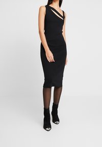 Abito midi aderente nero con un collo asimmetrico unico caratterizzato da dettagli intagliati. Il materiale è liscio e abbinato a stivaletti neri in suede alla caviglia.