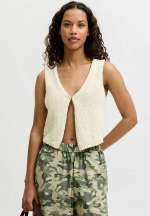 Giovane donna con capelli ricci che indossa un gilet crema senza maniche con texture e pantaloni verdi con fantasia mimetica, in piedi davanti a uno sfondo semplice.