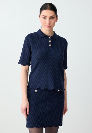 Frau trägt ein marineblaues, strukturiertes Poloshirt mit kurzen Ärmeln und einen passenden Rock mit gewellten Kanten und goldenen Knopfdetails.