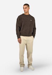 Brązowy puszysty sweter z okrągłym dekoltem, połączony z beżowymi zwężającymi się spodniami i białymi trampkami. Sweter ma małe logo na piersi.