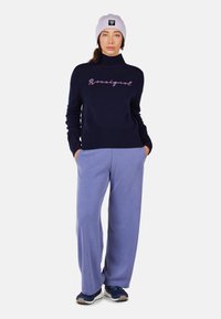 Navy knitted sweater with high collar and pink embroidered text; paired with light purple sweatpants and navy sports shoes. Light beanie worn.