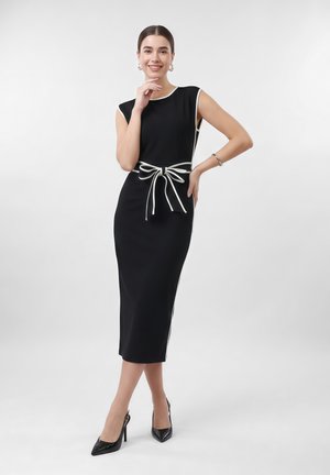 Žena v čiernych midi šatách bez rukávov s bielym lemovaním a viazanou mašľou v páse, v čiernych vysokých podpätkoch, usmievajúca sa s rukou pri tvári.