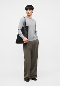 Mladý muž v šedém svetru a širokých hnědých kalhotách drží na rameni černou tašku a dívá se stranou.