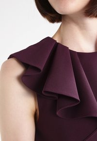 Detailní záběr na osobu v bezramínkových tmavě fialových šatech s několikavrstvými volány v oblasti ramen a výstřihu.