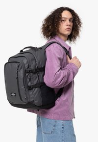 Zaino in nylon nero e grigio con numerosi scomparti con zip, spallacci imbottiti e decorazioni riflettenti. Indossato sulla schiena di un modello.