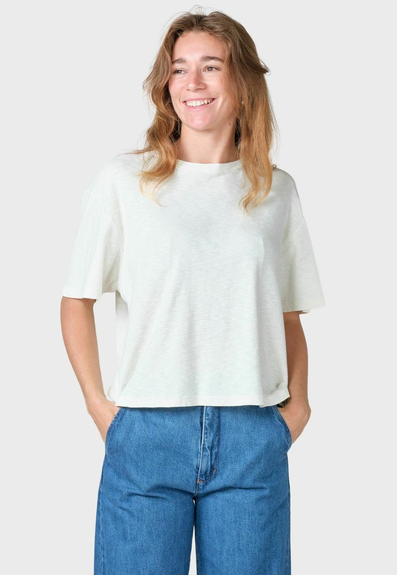 Jeune femme aux longs cheveux ondulés portant une chemise blanche ample à manches courtes et un jean bleu, souriante, les mains dans les poches.