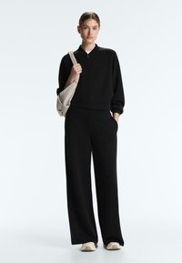 Femeie în picioare, purtând o jachetă neagră cu fermoar, pantaloni largi negri, adidași albi și o geantă bej pe umăr.