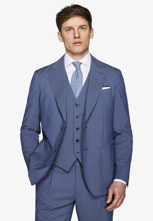 Mężczyzna ubrany w niebieski garnitur trzyczęściowy z białą koszulą i niebieskim krawatem w wzory, stojący na jednolitym białym tle.