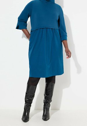 Femme portant une robe bleu sarcelle longueur genoux avec des manches trois-quarts, des collants noirs et des bottes en cuir noires montant jusqu'aux genoux, debout contre un mur blanc.
