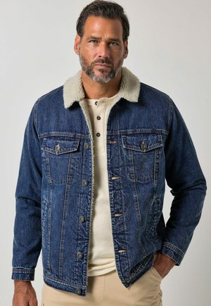 Mann mit Bart trägt blaue Jeansjacke mit weißem Fleece-Kragen sowie beiges Hemd und Hose, stehend vor schlichtem Hintergrund.