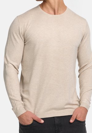 Homme portant un pull beige à manches longues et col rond, les mains dans les poches d'un pantalon noir, sur fond blanc.