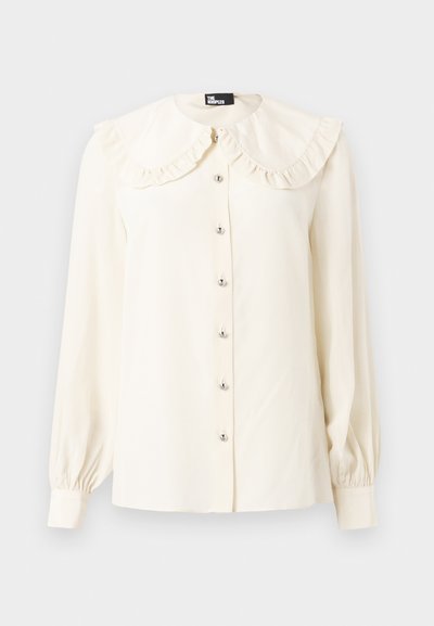 Κρεμ μπλούζα με μακριά μανίκια, μεγάλο βολάν γιακά και έξι ασημένια κουμπιά μπροστά, με μανσέτες που σφίγγουν και ευθεία βάση.