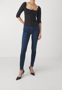 Top negro ajustado con escote cuadrado y cordón, combinado con vaqueros skinny azul oscuro y tacones negros puntiagudos con detalles verdes.