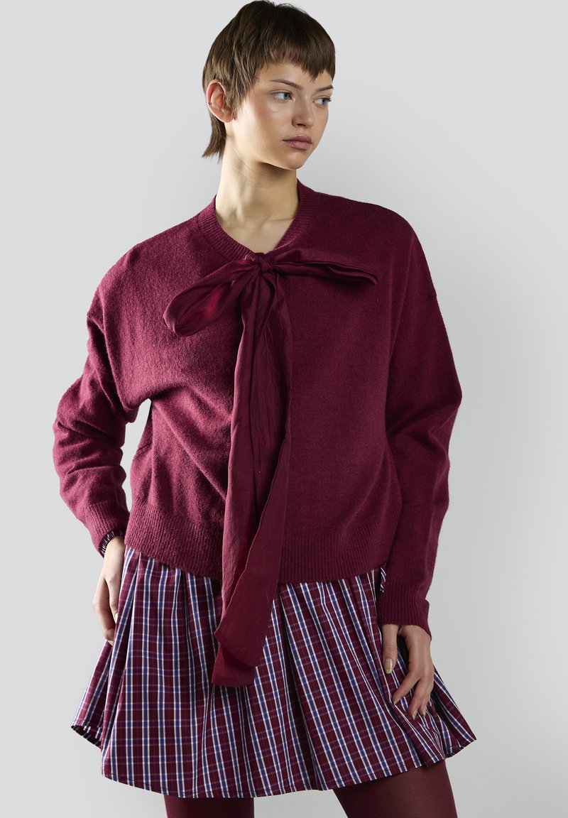 Maglione a maglia bordeaux con legatura frontale, abbinato a una gonna plissettata a quadrati nei colori bordeaux, blu e bianco, che mette in mostra texture sovrapposte.