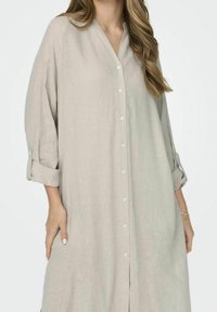 Abito camicia in lino beige chiaro con scollo a V, chiusura a bottoni frontale e maniche arrotolate. Presenta una vestibilità morbida e tasche laterali per una maggiore praticità.