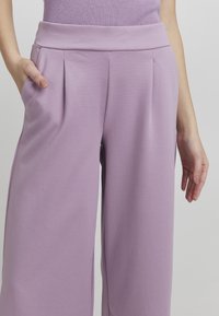 Liliowe spodnie o szerokich nogawkach wykonane z miękkiego materiału, z wysokim stanem, z fałdami z przodu oraz kieszeniami bocznymi. Gładka tekstura i luźny krój.