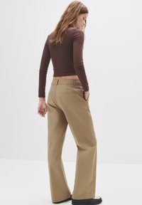 Beige weite Hose mit flacher Vorderseite, Gesäßtaschen und glattem Stoff. Kombiniert mit einem braunen, enganliegenden Langarmshirt.