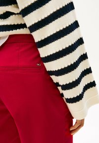 Un pull en maille rayé crème et marine associé à un pantalon rouge lisse. Les détails notables incluent une poche au dos du pantalon et des rayures texturées.