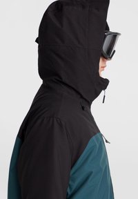 Chaqueta aislante negra y verde azulado con capucha ajustable, que presenta una textura exterior suave y un bolsillo con cremallera en el costado.