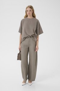 Νεαρή γυναίκα που φοράει φαρδιά καφέ ανοιχτή μπλούζα, παντελόνι φαρδιάς γραμμής ταιριαστό, λευκά τακούνια, κρατώντας μικρή καφέ τσάντα χειρός σε λευκό φόντο.