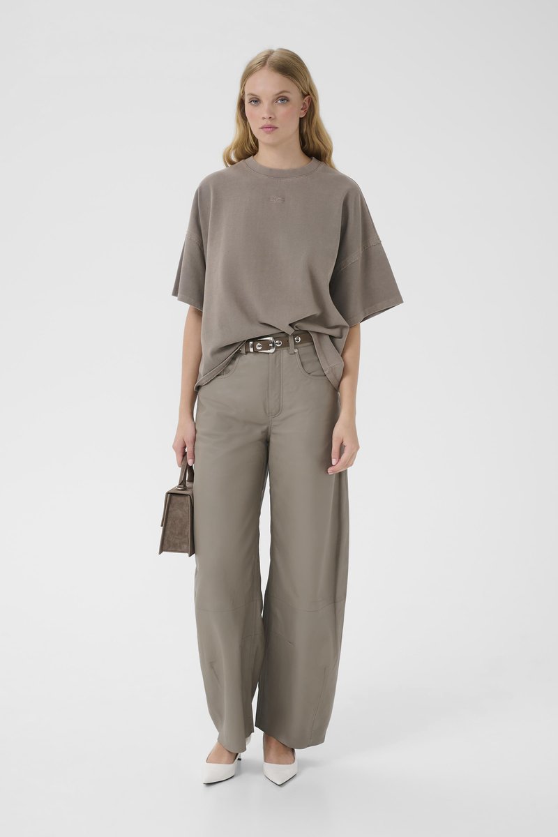 Νεαρή γυναίκα που φοράει φαρδιά καφέ ανοιχτή μπλούζα, παντελόνι φαρδιάς γραμμής ταιριαστό, λευκά τακούνια, κρατώντας μικρή καφέ τσάντα χειρός σε λευκό φόντο.